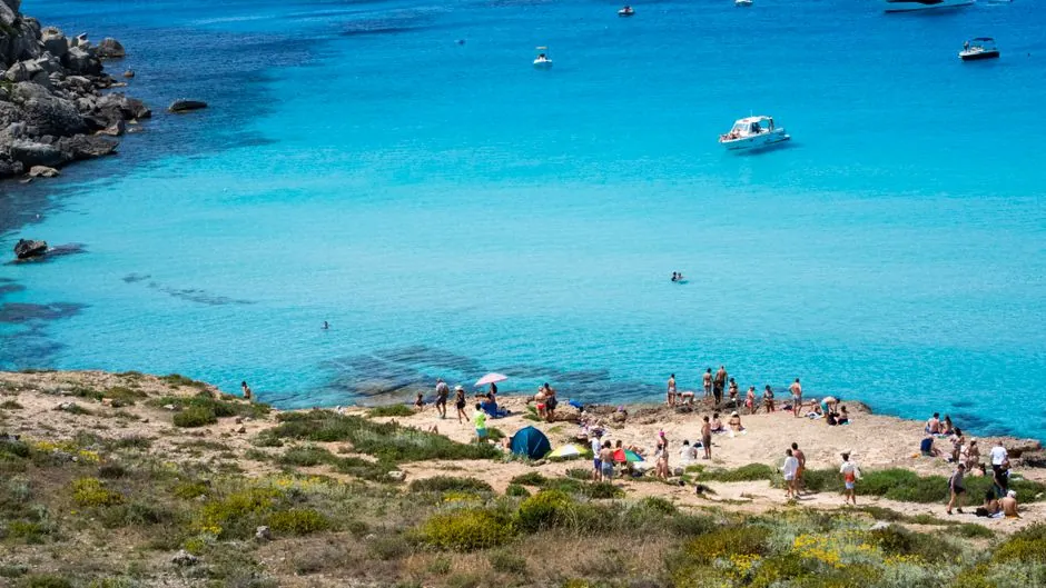Aerial view of Calamosche Beach, Sicily. Turquoise waters, boats, and beachgoers create a vibrant summer scene.