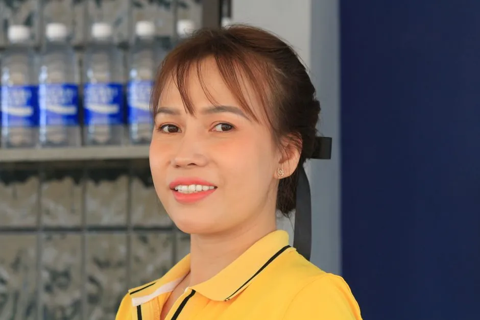 A smiling woman wearing a yellow shirt poses in front of a shelf with water bottles.