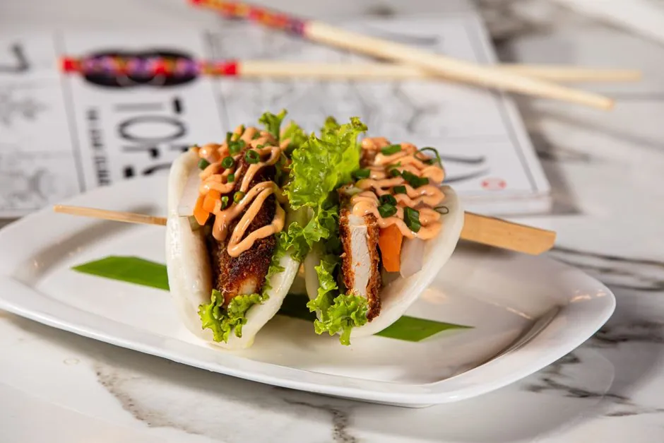 Tasty bao buns filled with crispy pork, lettuce, and sauce on a marble table.