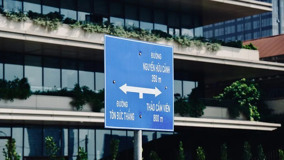 Blue street sign directing traffic amidst modern urban architecture and greenery.