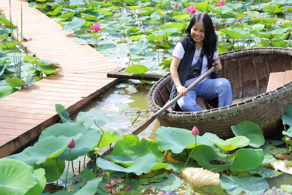 Smiling woman rowing a traditional basket boat through a vibrant lotus pond under sunny skies.