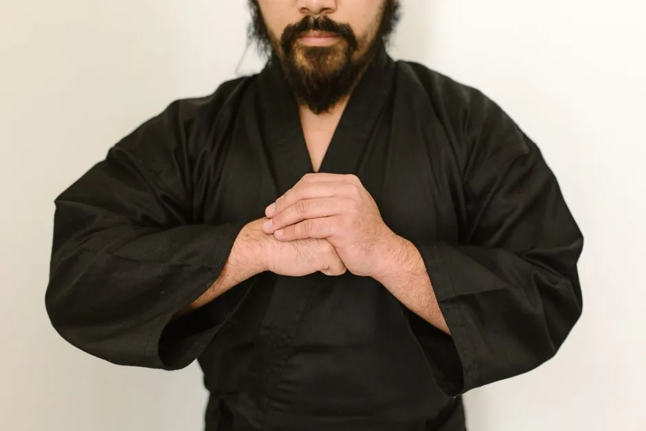 Bearded martial artist in black gi poised with a hand gesture, embodying focus and discipline indoors.